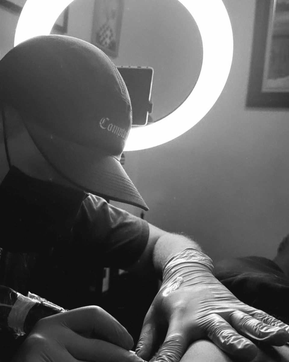 Davi Pimentel tatuando, de boné e luvas, com uma luz circular ao fundo, em preto e branco